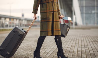 Pasageră cu troler și pașaport în fața unui aeroport, ilustrând scumpirea biletelor de avion