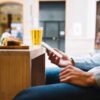 Rezervări hoteluri Vola: bărbat care folosește telefonul la o terasă, cu bere și o gustare pe masă