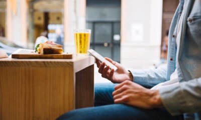 Rezervări hoteluri Vola: bărbat care folosește telefonul la o terasă, cu bere și o gustare pe masă